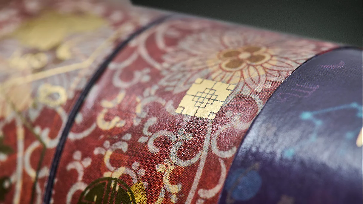 Close-up of a textured, patterned packaging with red and purple colors and gold foil accents, featuring floral and decorative designs and the KURZ logo.