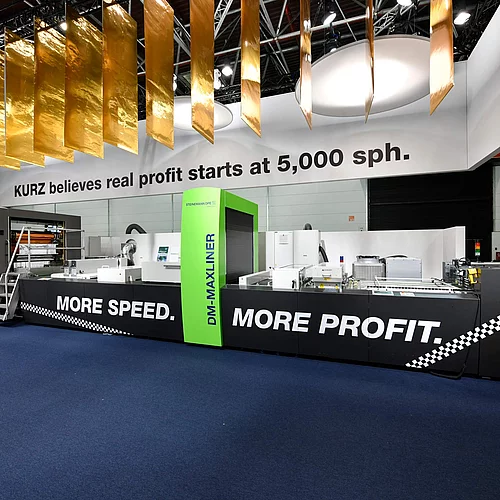 KURZ trade show booth showcasing the DM-MAXLINER digital embellishment machine with “More Speed. More Profit.” branding and hanging gold foil decorations.