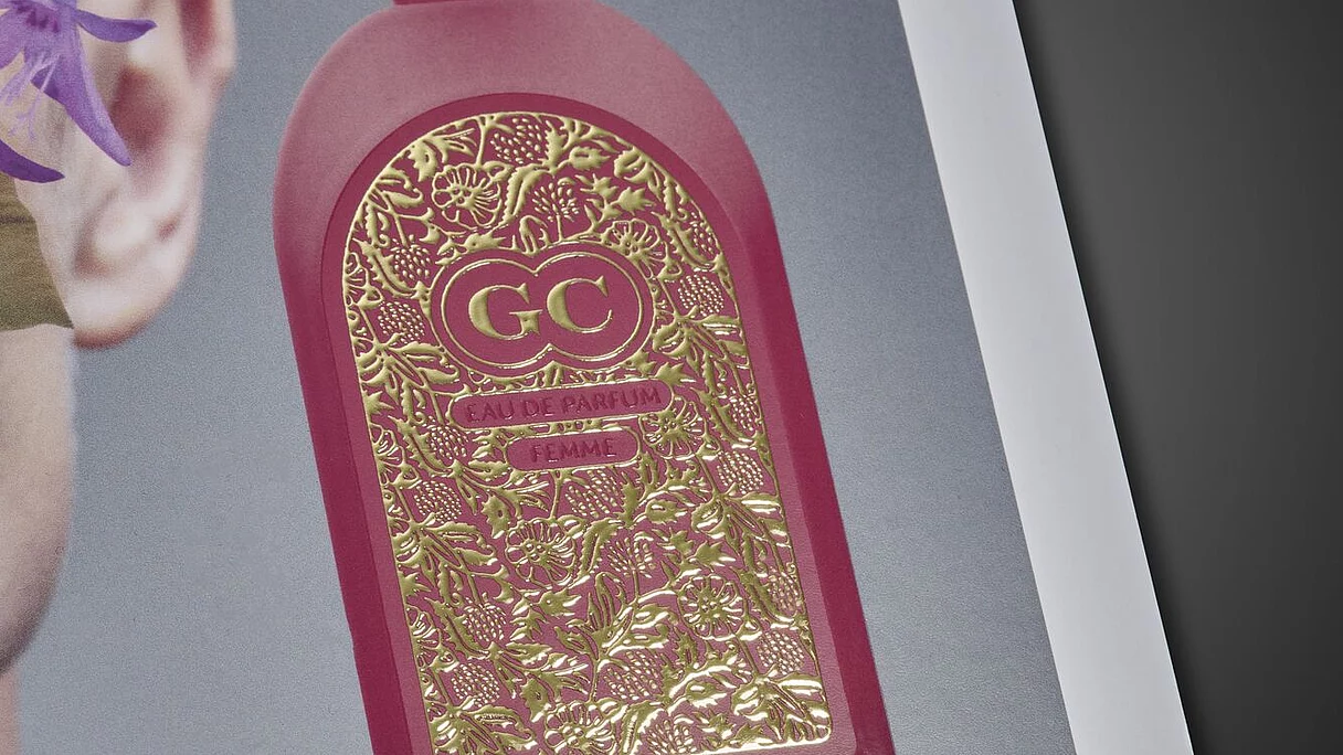 Close-up of a magazine cover featuring a red perfume bottle with intricate gold foil floral design, and embossed "STEINEMANN DPE" branding at the bottom.