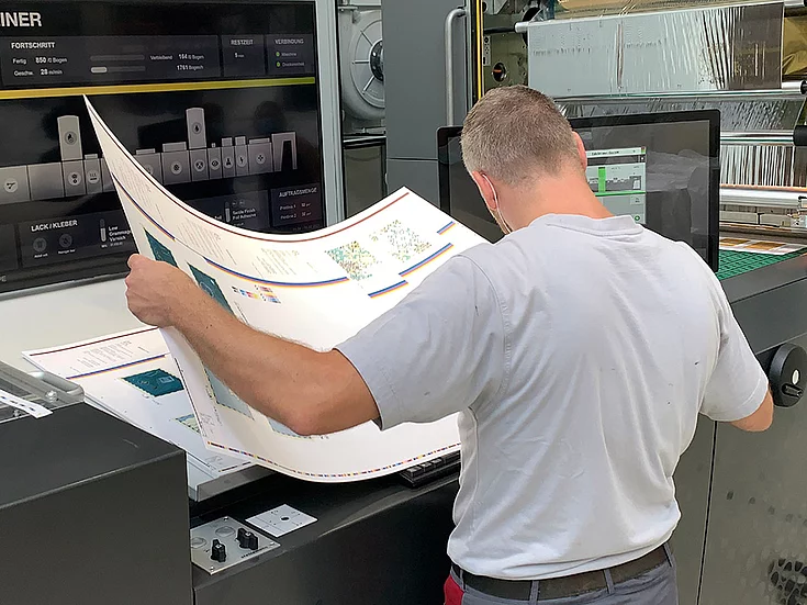 Man in white shirt inspecting printed sheets in a printing facility, surrounded by Maxliner 3D Printer machines and computer monitors.
