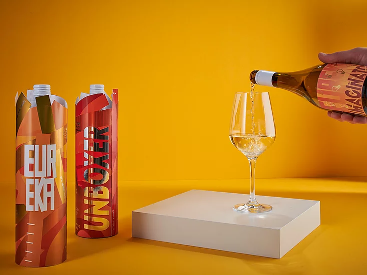 Hand pouring white wine from a bottle labeled ‘IMAGINATION’ into a glass on a white pedestal, with two colorful wine bottles in decorative sleeves on a yellow background.