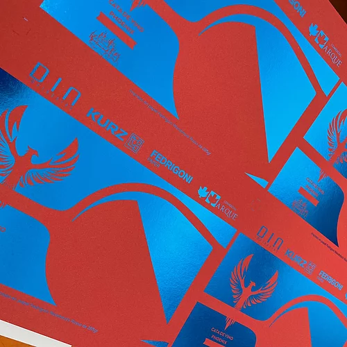 Close-up of red printed sheet with vibrant blue foil accents, featuring a phoenix logo and text including "CATA DE VINO PHOENIX," "D.I.N. impresores," "KURZ," "FEDRIGONI PAPER," and "COMERCIAL ARQUE," showcasing advanced print and foil techniques.