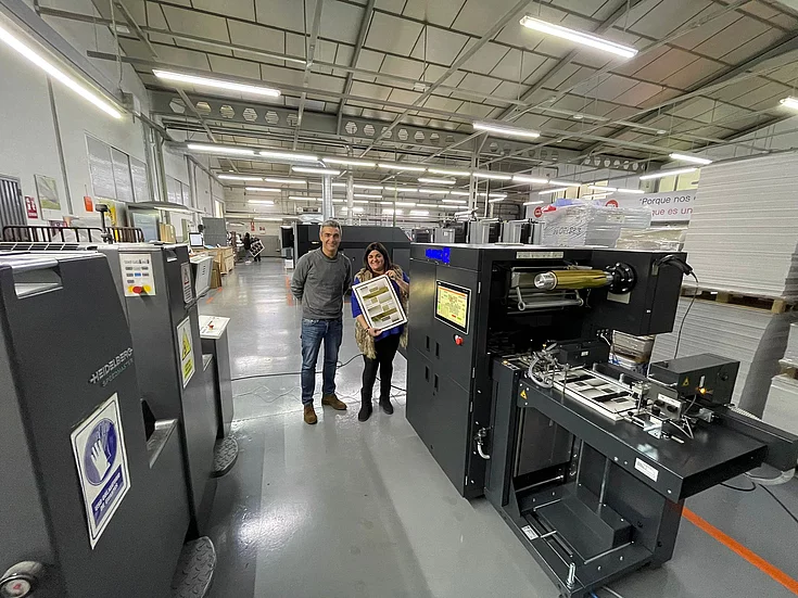 Two people standing in a large industrial printing facility, smiling and holding a printed sheet with gold foil details, surrounded by heavy printing machinery and stacks of paper.