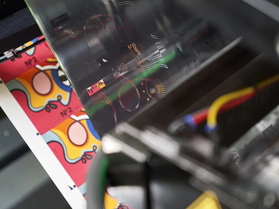 Close-up of KURZ digital embellishment machine applying holographic foil to colorful printed sheet with abstract artwork and product label design. 