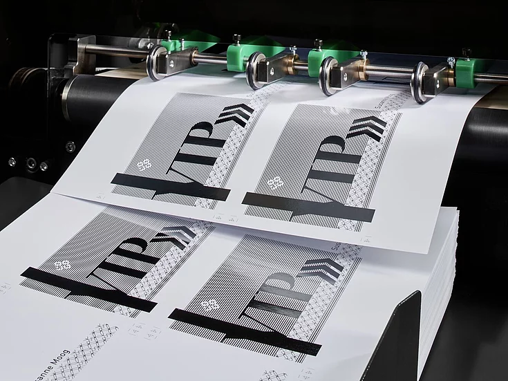 Close-up of a printing machine producing sheets with a design labeled ‘LUXORO’ featuring intricate line patterns and holographic elements.
