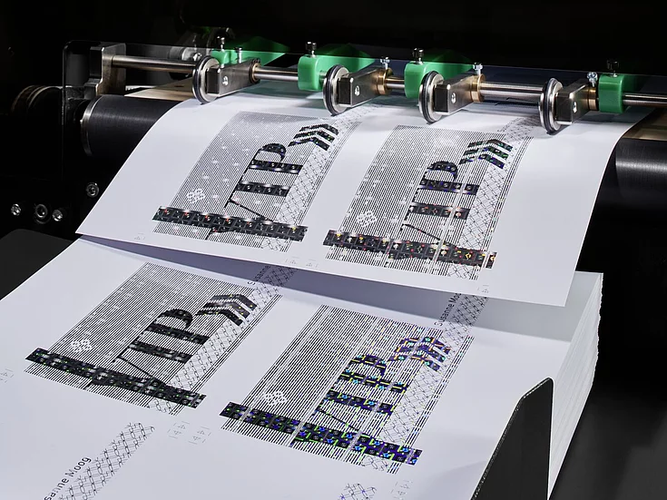 Nahaufnahme einer Druckmaschine, die Blätter mit einem Design namens „LUXORO“ produziert, das sich durch komplexe Linienmuster und holografische Elemente auszeichnet.
