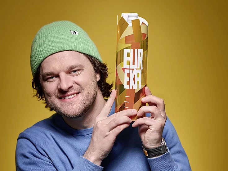 Man with a green knit hat and blue sweater smiles while holding a tall, orange patterned carton with the word "EUREKA" in bold white letters, against a solid yellow background.