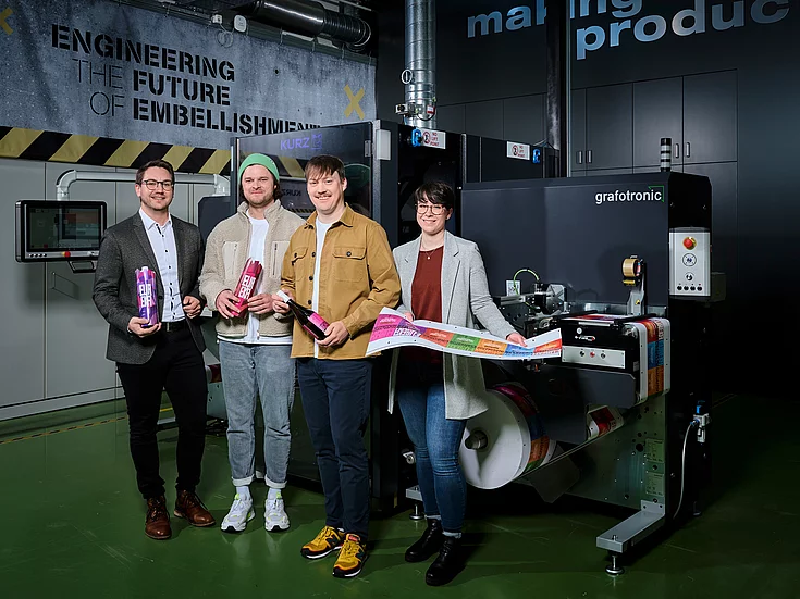 Group portrait of four people standing in a factory setting beside a large "grafotronic" machine, holding colorful packaging and a printed sheet with vibrant designs. Wall text reads "ENGINEERING THE FUTURE OF EMBELLISHMENT."