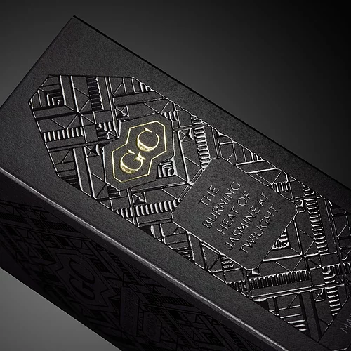  Luxurious black packaging with intricate embossed geometric patterns, gold foil "GC" logo, and silver debossed text reading "The burning heat of jasmine at twilight" and "Made in France."