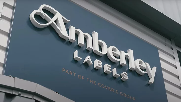 Outdoor signage on a building with the text "Amberley Labels" in raised white letters, and smaller text below reading "Part of the Coveris Group" in copper foil on a blue background.