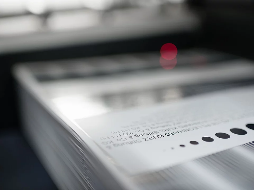 Close-up image showing a stack of printed sheets with a glossy, smooth finish on the top sheet. The text and barcode details are partially visible, and there is a soft, out-of-focus red light reflection in the background