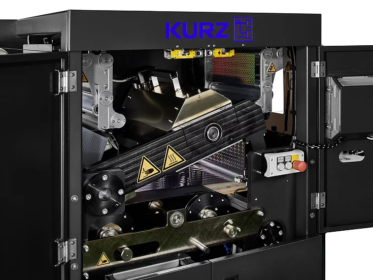 Close-up of open KURZ foiling machine showing rollers, heating elements, and control switches with warning labels, highlighting intricate industrial components and advanced foil application technology.