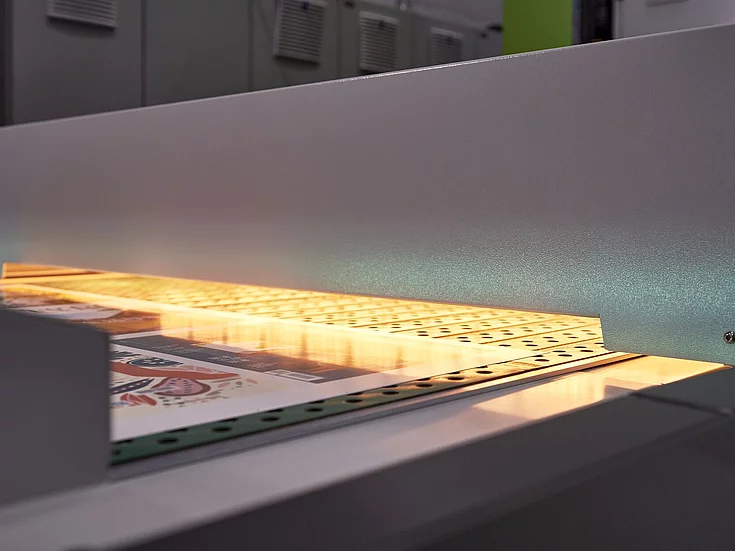 Close-up of a printing machine scanning or inspecting colorful printed sheets moving on a conveyor belt under a bright light.