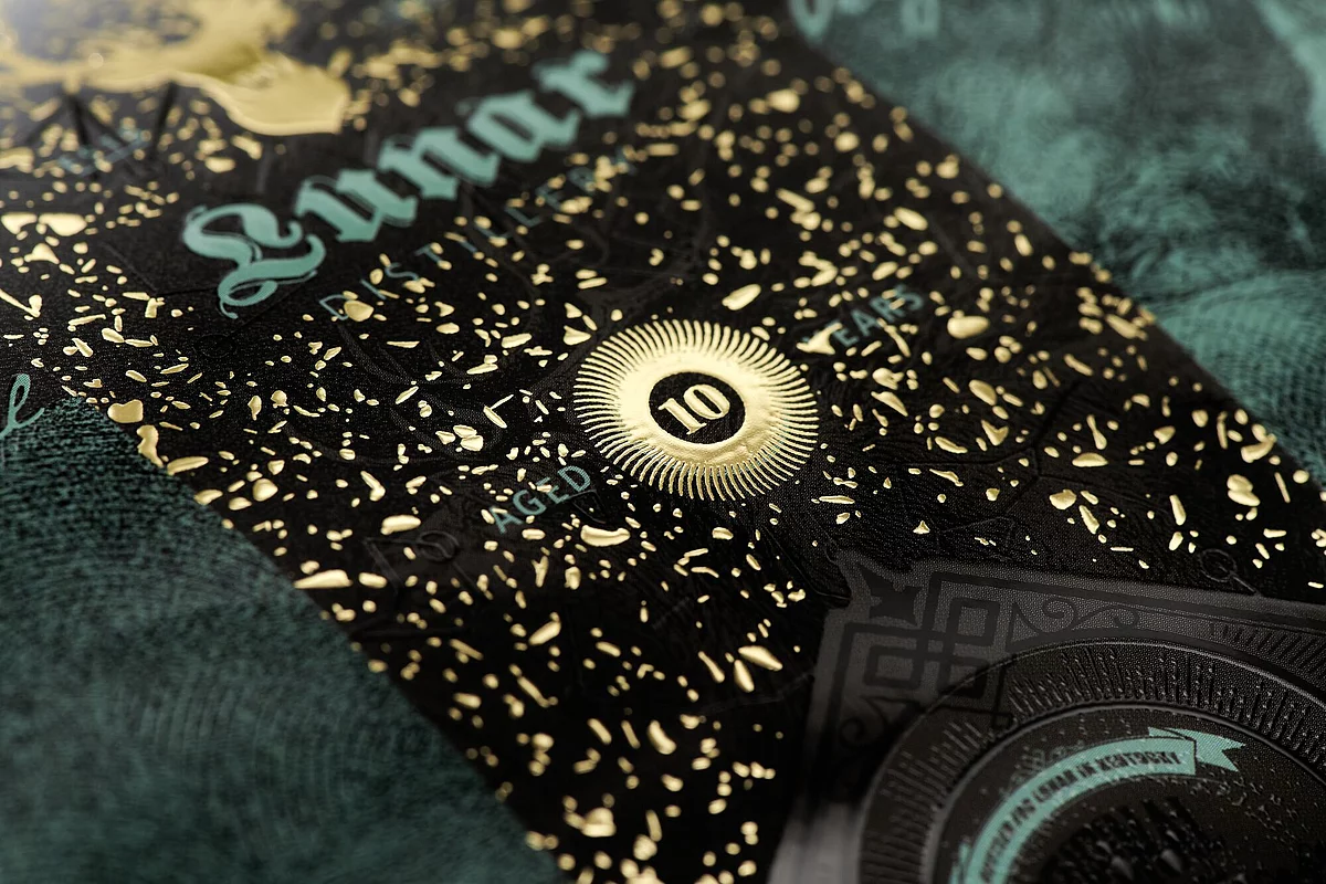 Close-up of a black and teal textured label with intricate gold foil detailing, featuring the text "AGED 10 YEARS" in the center, showcasing luxury print effects.
