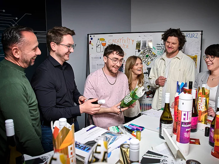 Group of six young adults gathered around a table, smiling and examining colorful packaging designs in a creative office setting.