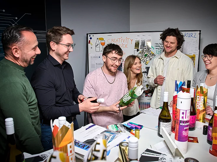 Group of six young adults gathered around a table, smiling and examining colorful packaging designs in a creative office setting.