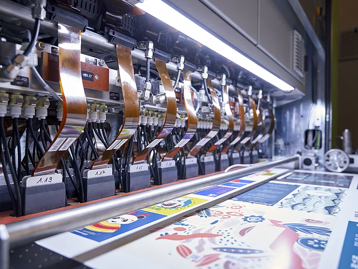 Close-up of an industrial digital printing machine in operation, with colorful printed sheets coming out and multiple copper-colored ribbon cables connected to the printheads.