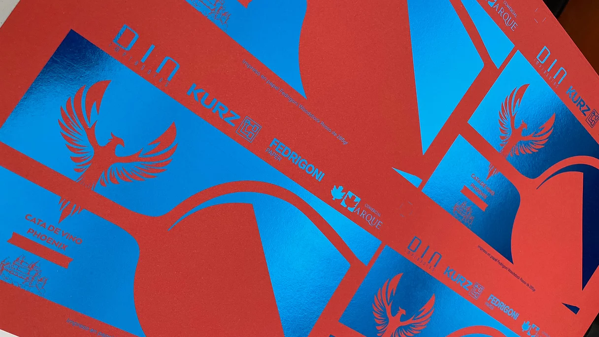 Close-up of red printed sheet with vibrant blue foil accents, featuring a phoenix logo and text including "CATA DE VINO PHOENIX," "D.I.N. impresores," "KURZ," "FEDRIGONI PAPER," and "COMERCIAL ARQUE," showcasing advanced print and foil techniques.