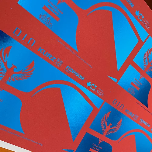 Close-up of red printed sheet with vibrant blue foil accents, featuring a phoenix logo and text including "CATA DE VINO PHOENIX," "D.I.N. impresores," "KURZ," "FEDRIGONI PAPER," and "COMERCIAL ARQUE," showcasing advanced print and foil techniques.