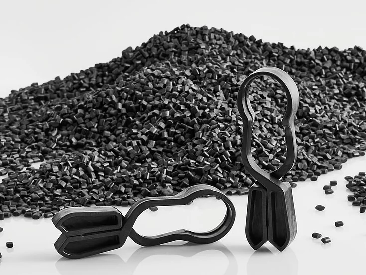 Two black plastic clips stand upright next to a pile of small black plastic pellets on a reflective white surface.