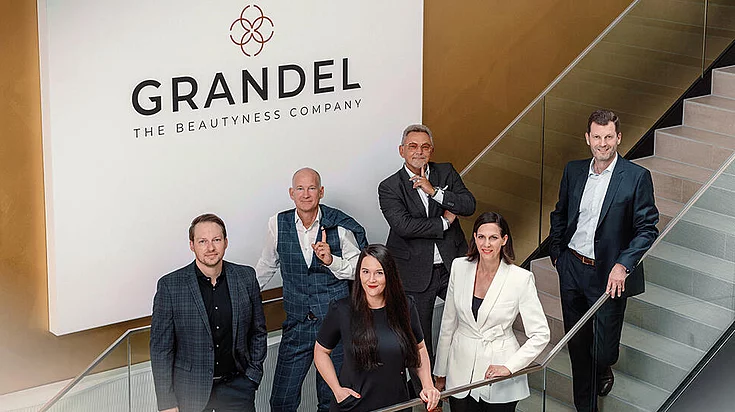 Gruppenfoto von sechs professionell gekleideten Personen, die auf einer Treppe unter einem Schild mit der Aufschrift „GRANDEL THE BEAUTYNESS COMPANY” in einem modernen Bürogebäude posieren.