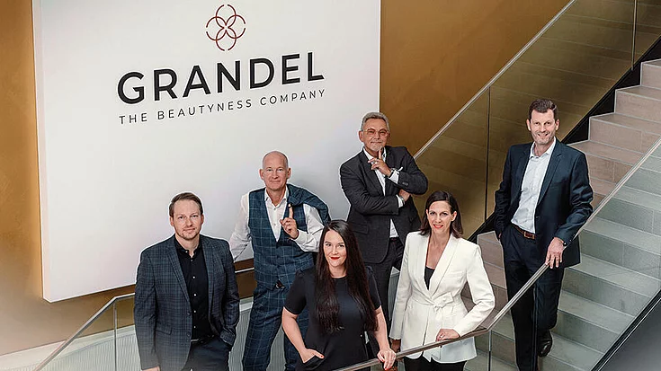Gruppenfoto von sechs Personen in Businesskleidung auf einer Treppe unter dem Schild „GRANDEL THE BEAUTYNESS COMPANY“.