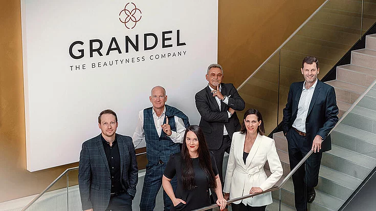 Group photo of six professionally dressed people posing on a staircase under a sign that reads "GRANDEL THE BEAUTYNESS COMPANY" inside a modern office building.