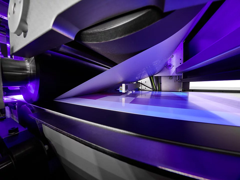 Close-up of a label printing machine applying glossy coating on printed sheets with purple light reflection.