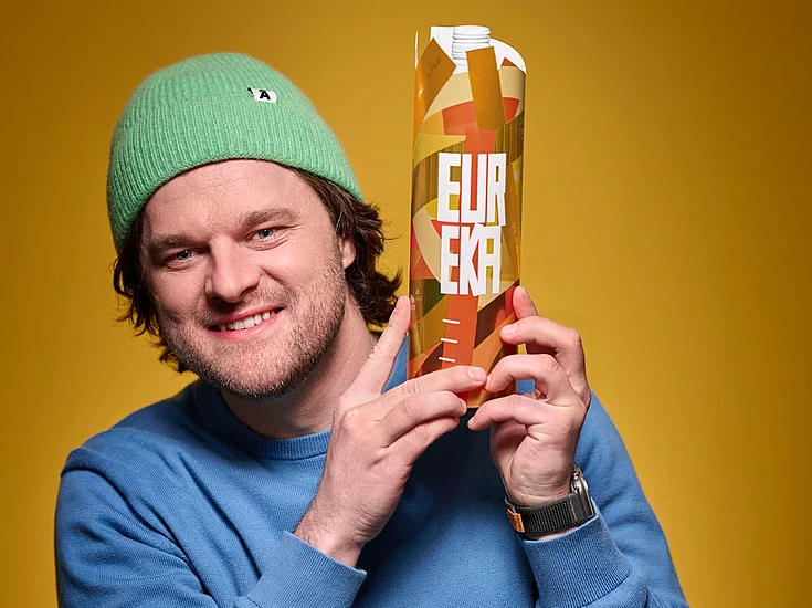 Ein Mann mit einer grünen Strickmütze und einem blauen Pullover lächelt, während er einen hohen Karton mit orangefarbenem Muster und dem Wort „EUREKA” in fetten weißen Buchstaben vor einem einfarbigen gelben Hintergrund hält.