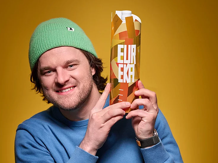 Man wearing green hat and blue sweater smiling while holding orange “EUREKA” carton against yellow background.