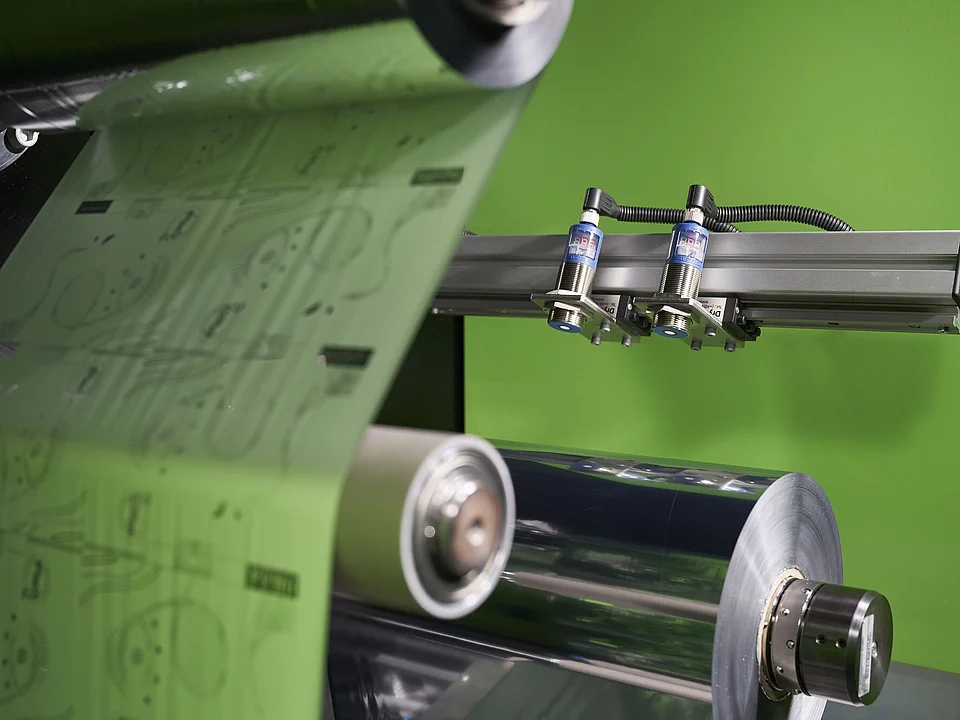 Close-up of a digital Embellishment Technologie showing transparent sheets with printed designs rolling on metal rollers, against a bright green background.