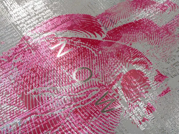 Close-up of a textured print with pink brush strokes and silver text forming the word ‘NOW’ on a metallic background.