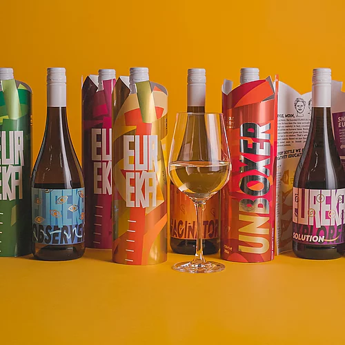 Six colorful wine bottles in decorative sleeves with bold text and designs, arranged in a row on a yellow background, with a glass of white wine in front.