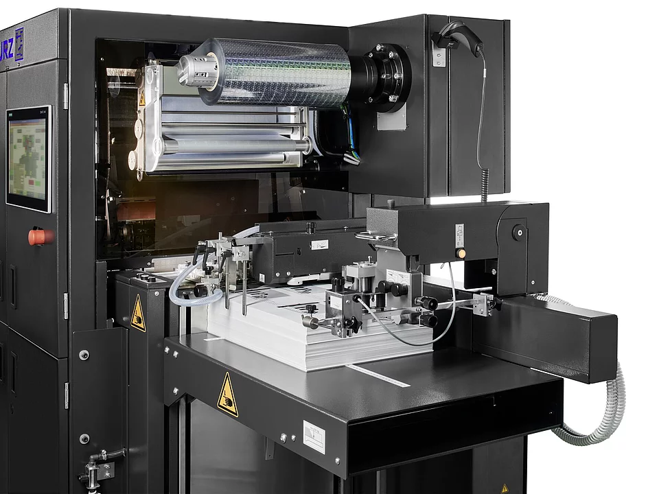  Digital embellishment machine by KURZ applying foil to stacked sheets, with visible foil roll, control arms, and touchscreen interface.