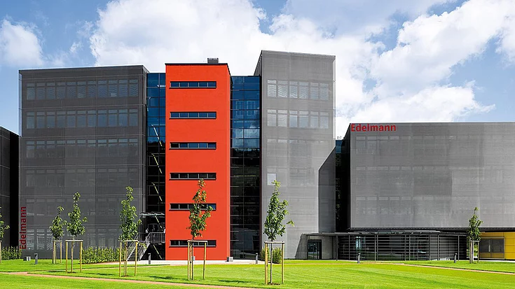 Modern office building with a red central section flanked by large glass and metal facades, surrounded by a neatly manicured lawn and young trees, with "Edelmann" signage visible on both sides.