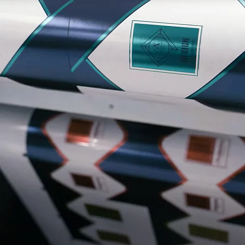 Printing press producing rolls of patterned labels with diamond-shaped frames, teal and white design on one roll, and copper and white on another, showing the design process in action.