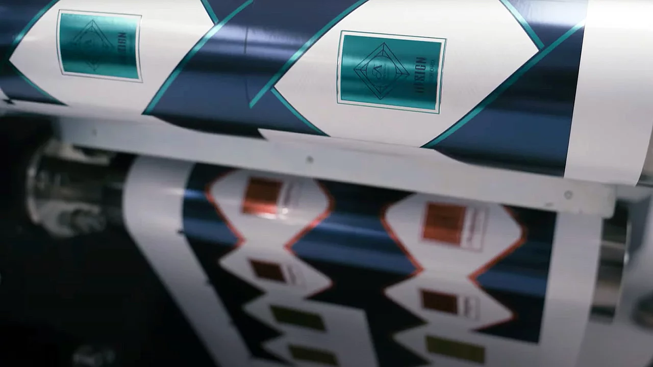 Printing press producing rolls of patterned labels with diamond-shaped frames, teal and white design on one roll, and copper and white on another, showing the design process in action.
