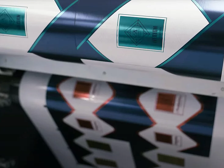 Printing press producing rolls of patterned labels with diamond-shaped frames, teal and white design on one roll, and copper and white on another, showing the design process in action.