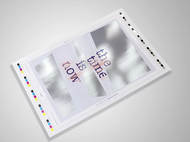 Printing proof sheet with metallic silver blocks and the phrase ‘the time is now’ in a typewriter-style font, viewed at an angle on a gray surface.