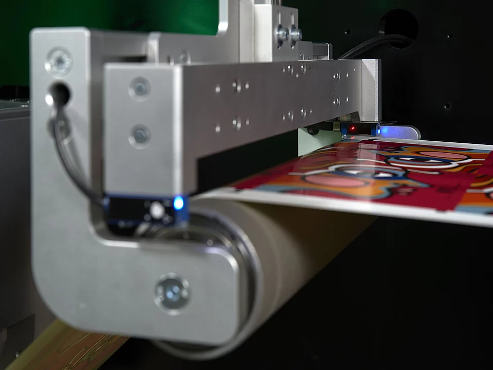 Close-up of a digital metallizing technologie, showing a vibrant red printed sheet moving through a roller system with metal components and sensors emitting blue and red lights.