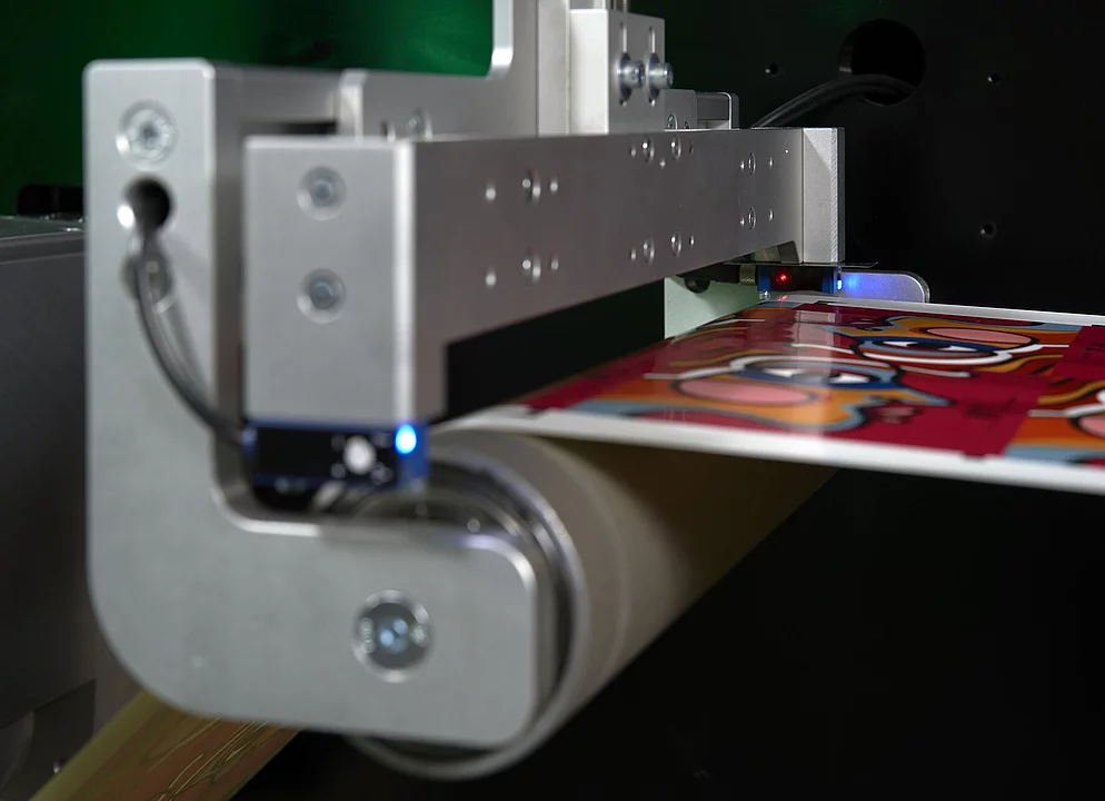 Nahaufnahme einer digitalen Metallisierungs-Technologie, die ein leuchtend rotes bedrucktes Blatt zeigt, das sich durch ein Rollensystem mit Metallkomponenten und Sensoren bewegt, die blaues und rotes Licht ausstrahlen.