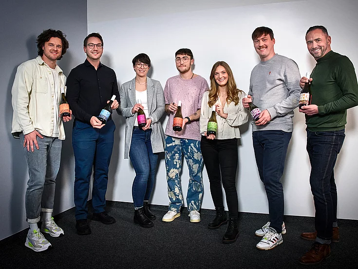 Group photo of seven people standing indoors, holding wine bottles with colorful labels. They appear cheerful and are posing for the camera. The background is plain, likely a studio or office setting.