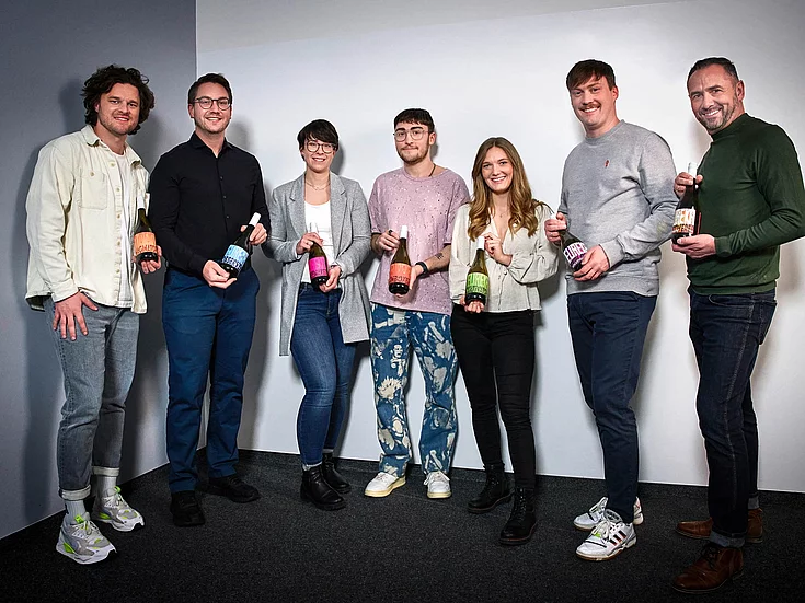 Group photo of seven people standing indoors, holding wine bottles with colorful labels. They appear cheerful and are posing for the camera. The background is plain, likely a studio or office setting.
