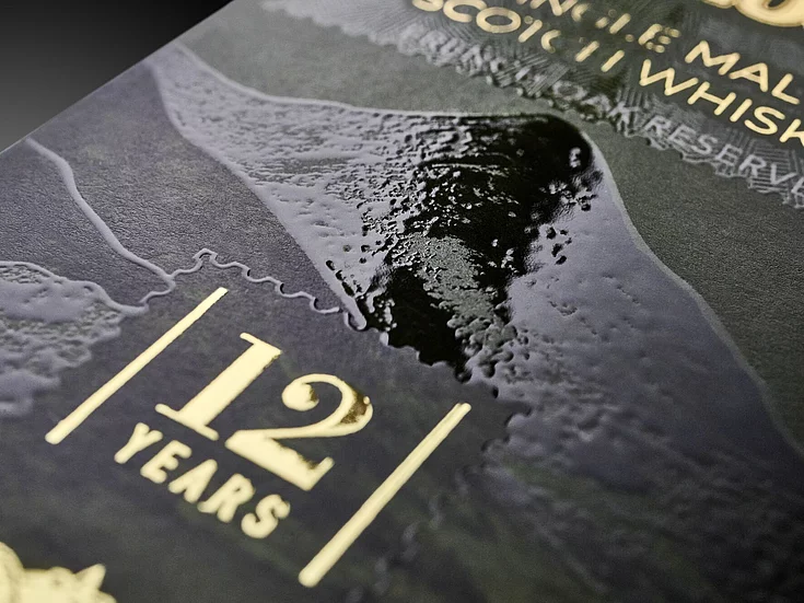 Close-up of premium whisky packaging with glossy spot varnish and gold foil embossing, highlighting “12 Years” age statement and textured landscape design.