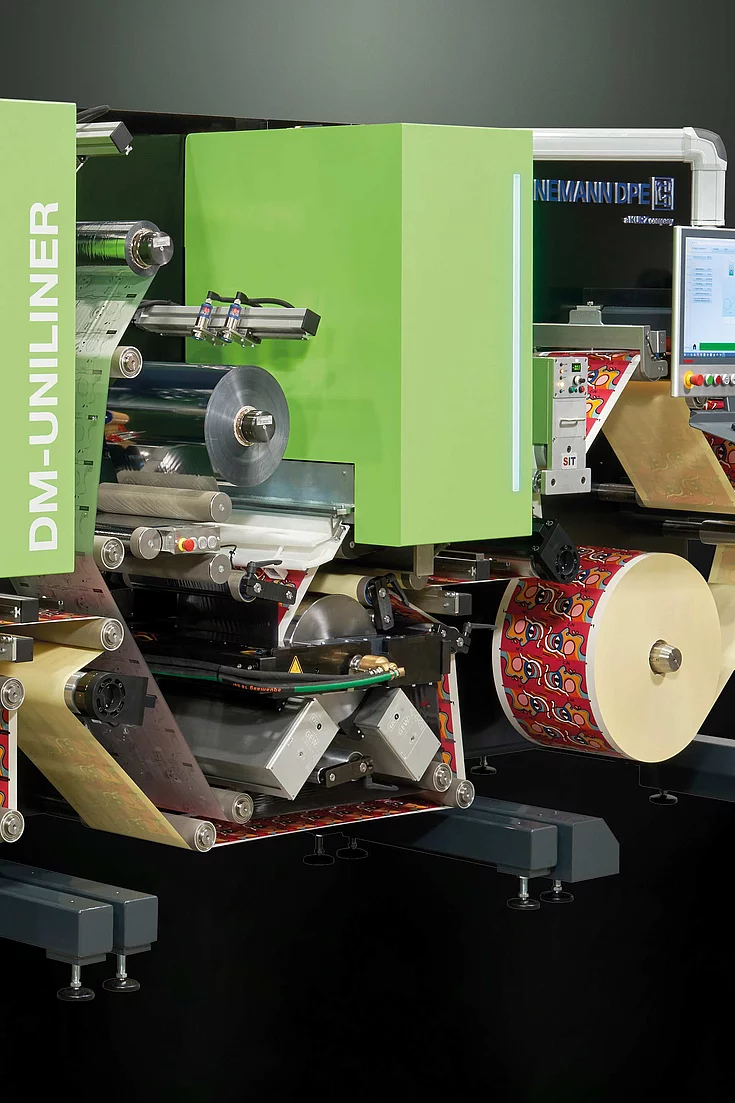 Digital Embellishment Solution with green panels, multiple rollers, two screens, and roll-fed colorful label material.