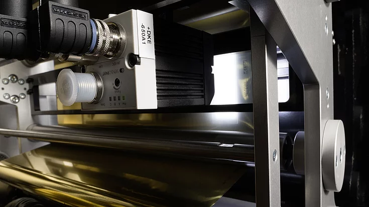 Close-up of printing machinery with metallic gold foil on a roll, showing intricate industrial components and a partially visible control panel.