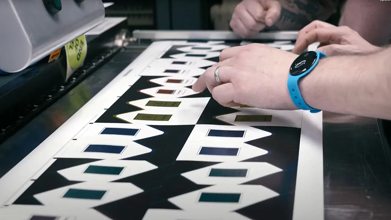 Close-up of hands inspecting printed sheets with geometric patterns and metallic gold foil accents, on a flatbed printer in a production setting.