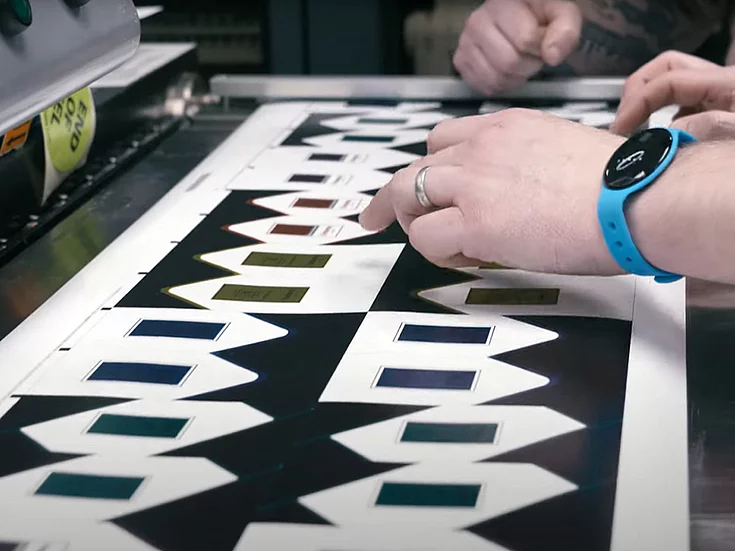 Close-up of hands inspecting printed sheets with geometric patterns and metallic gold foil accents, on a flatbed printer in a production setting.