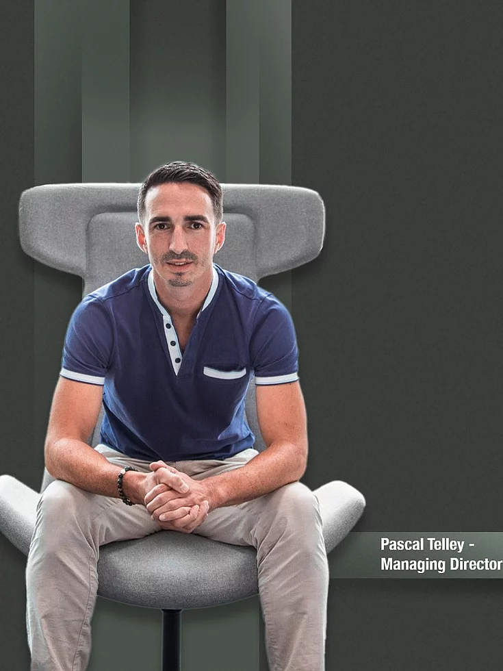 Man sitting relaxed on a gray designer chair against a dark background.