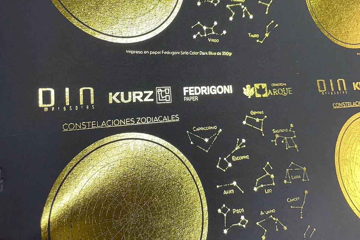 Close-up of black printed sheet with gold foil accents, showing zodiac constellations and logos for D.I.N. impresores, KURZ, FEDRIGONI Paper, and Comercial Arque, emphasizing premium print quality.